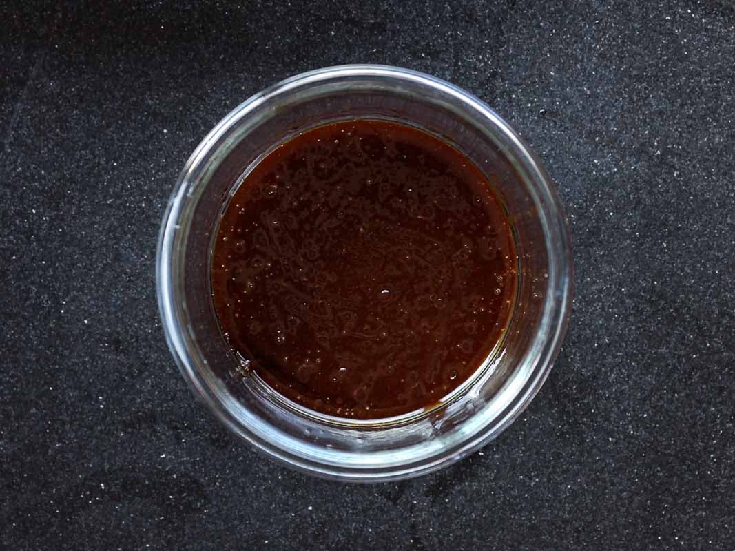 A glass bowl filled with dark brown sauce sits on a black textured countertop. The sauce appears thick and glossy, with a consistent texture throughout the bowl.